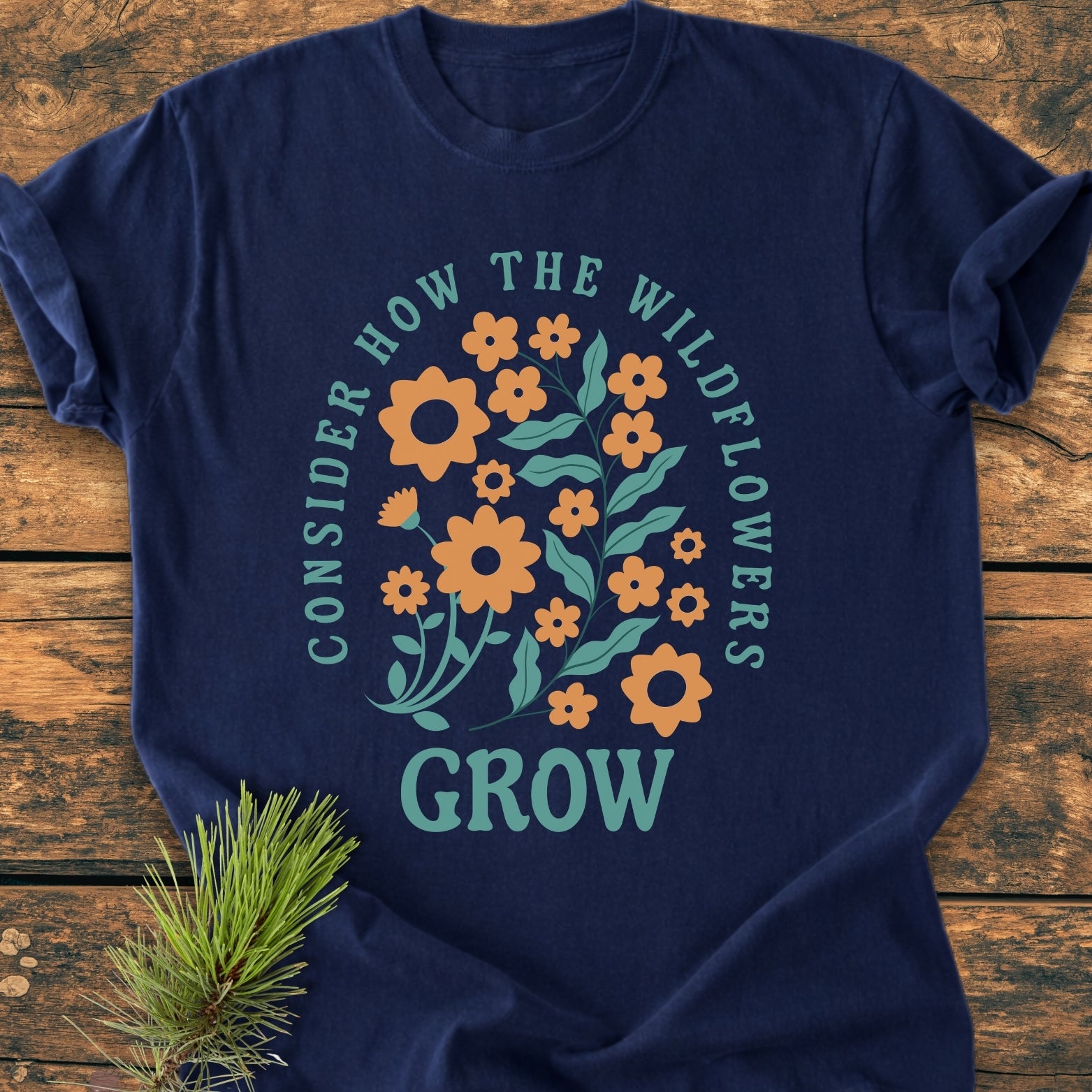 Wildflowers Luke 12,27 - Vintage Wash Tee features navy fabric with a stylized orange wildflower design and motivational text. Laid on rustic wood with an evergreen sprig for styling.