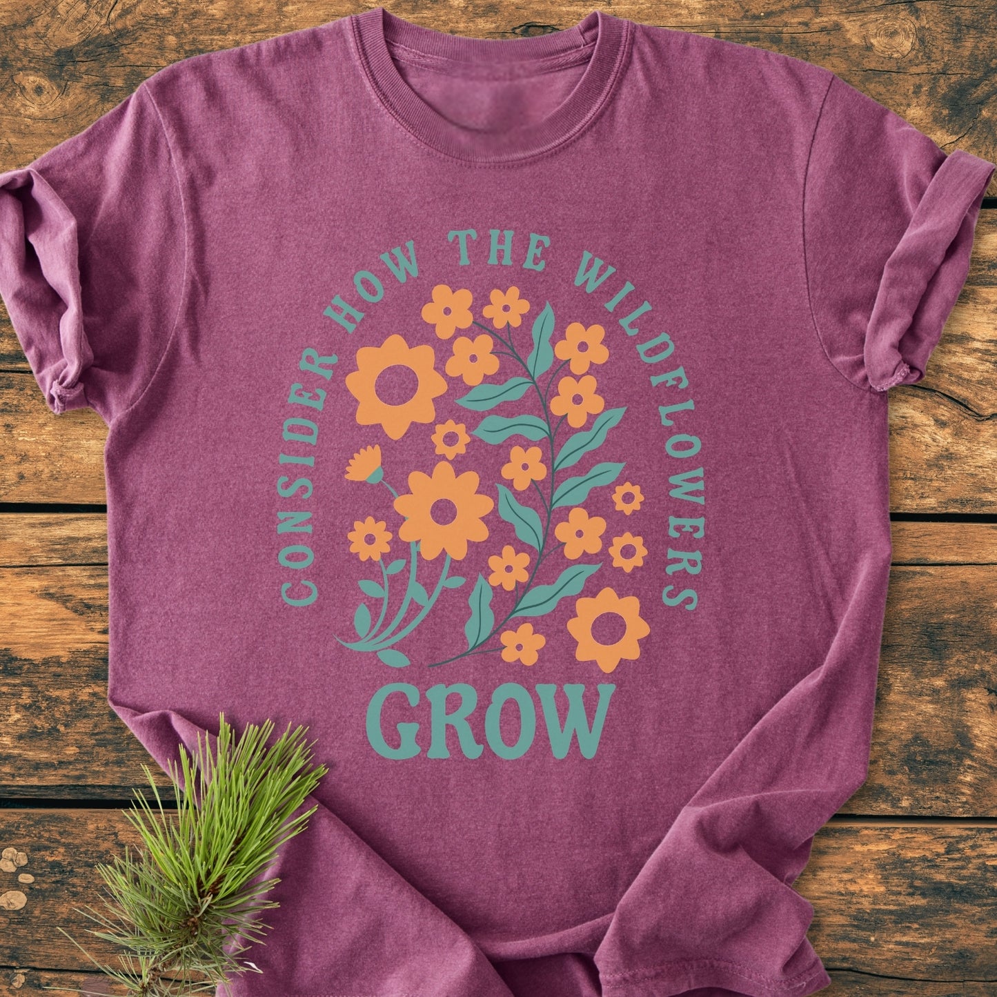 Wildflowers Luke 12,27 - Vintage Wash Tee features a graphic of orange wildflowers with teal leaves and the words “CONSIDER HOW THE WILDFLOWERS GROW” on a mauve T-shirt, laid on wood.