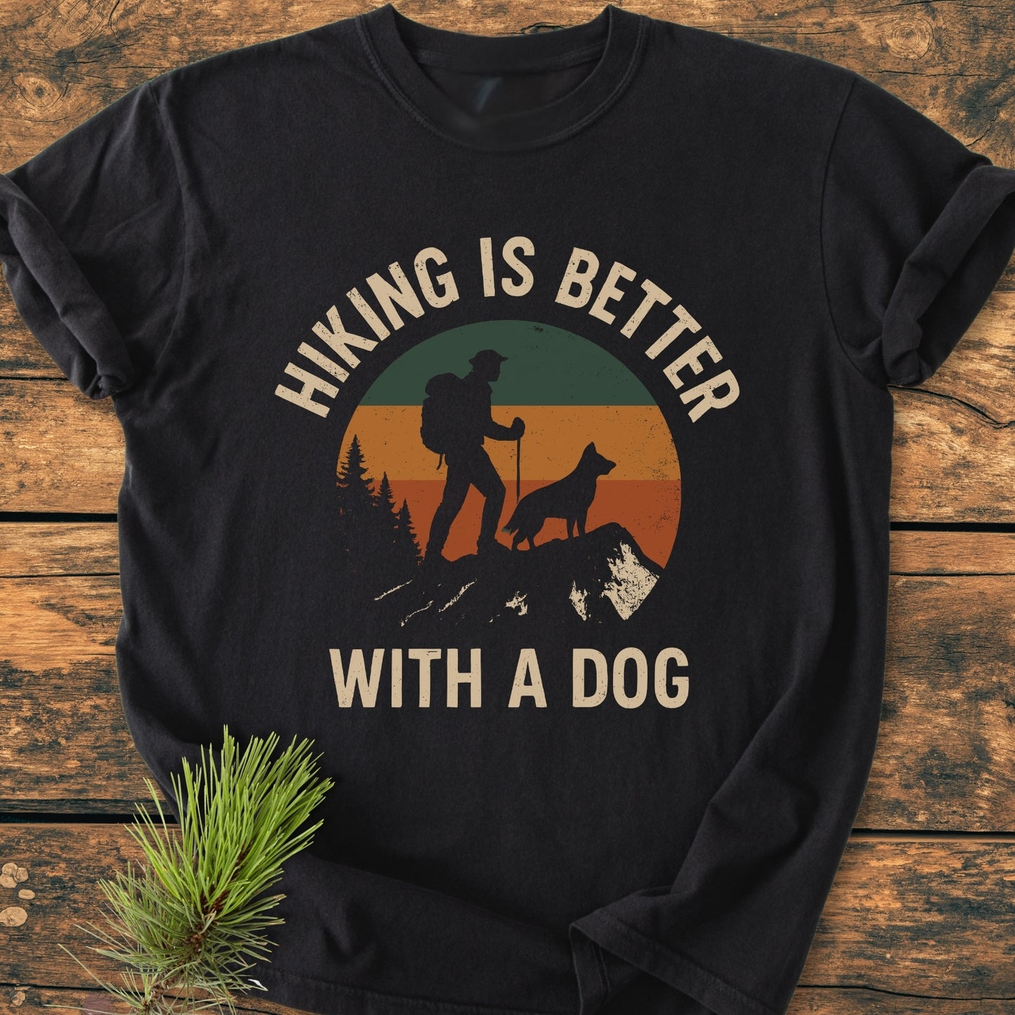 Hiking Is Better With A Dog - Vintage Wash Tee featuring a circular graphic of a hiker and dog silhouette against a sunset backdrop, laid flat on a wooden surface.