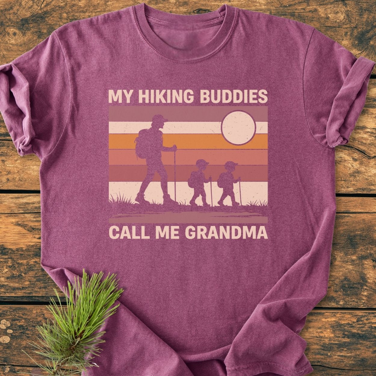 My Hiking Buddies Call Me Grandma - Vintage Wash Tee features a retro sunset graphic of a hiking grandma with kids. Includes a small pine twig accentuating its nature theme.