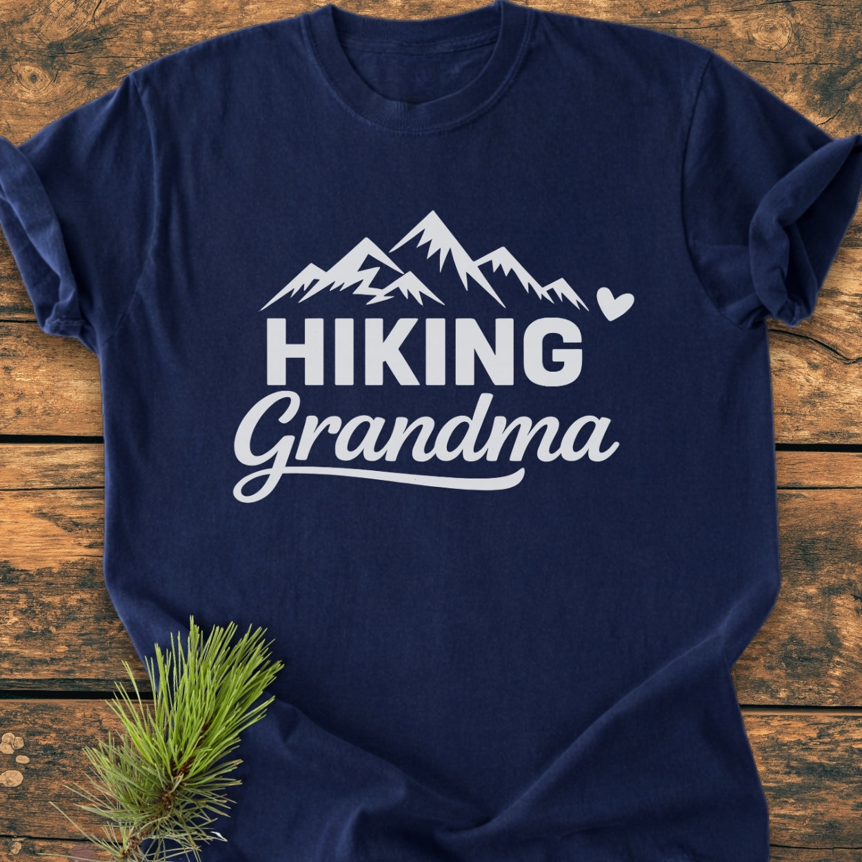 Navy-blue Hiking Grandma - Vintage Wash Tee with white mountain print, HIKING and Grandma text, and heart accent. Laid on wooden surface, featuring soft-washed, garment-dyed cotton for a relaxed fit.