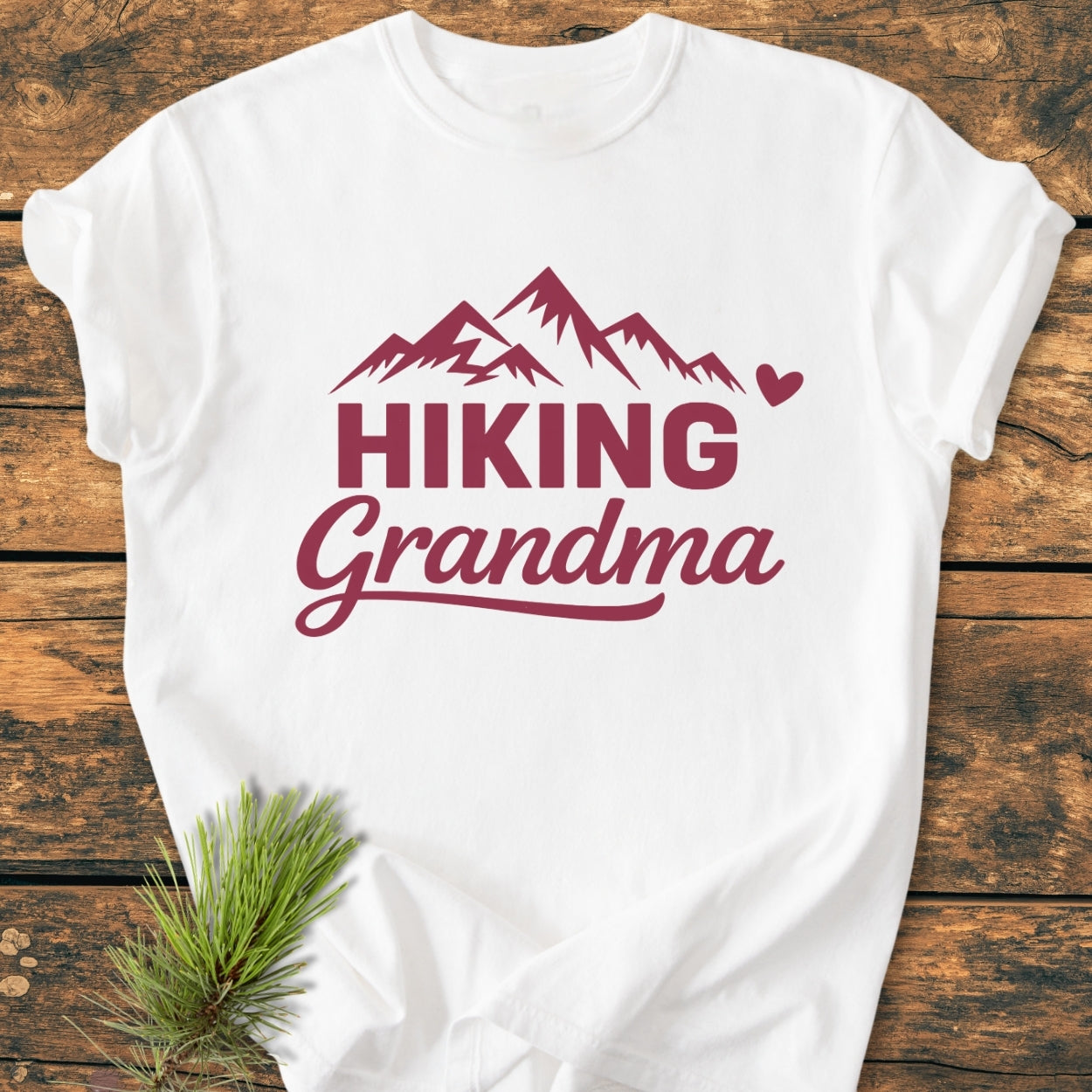 Hiking Grandma - Vintage Wash Tee: White T-shirt with stylized maroon mountain peaks and text, HIKING Grandma, accented by a heart graphic. Laid on wood, styled with pine greenery.