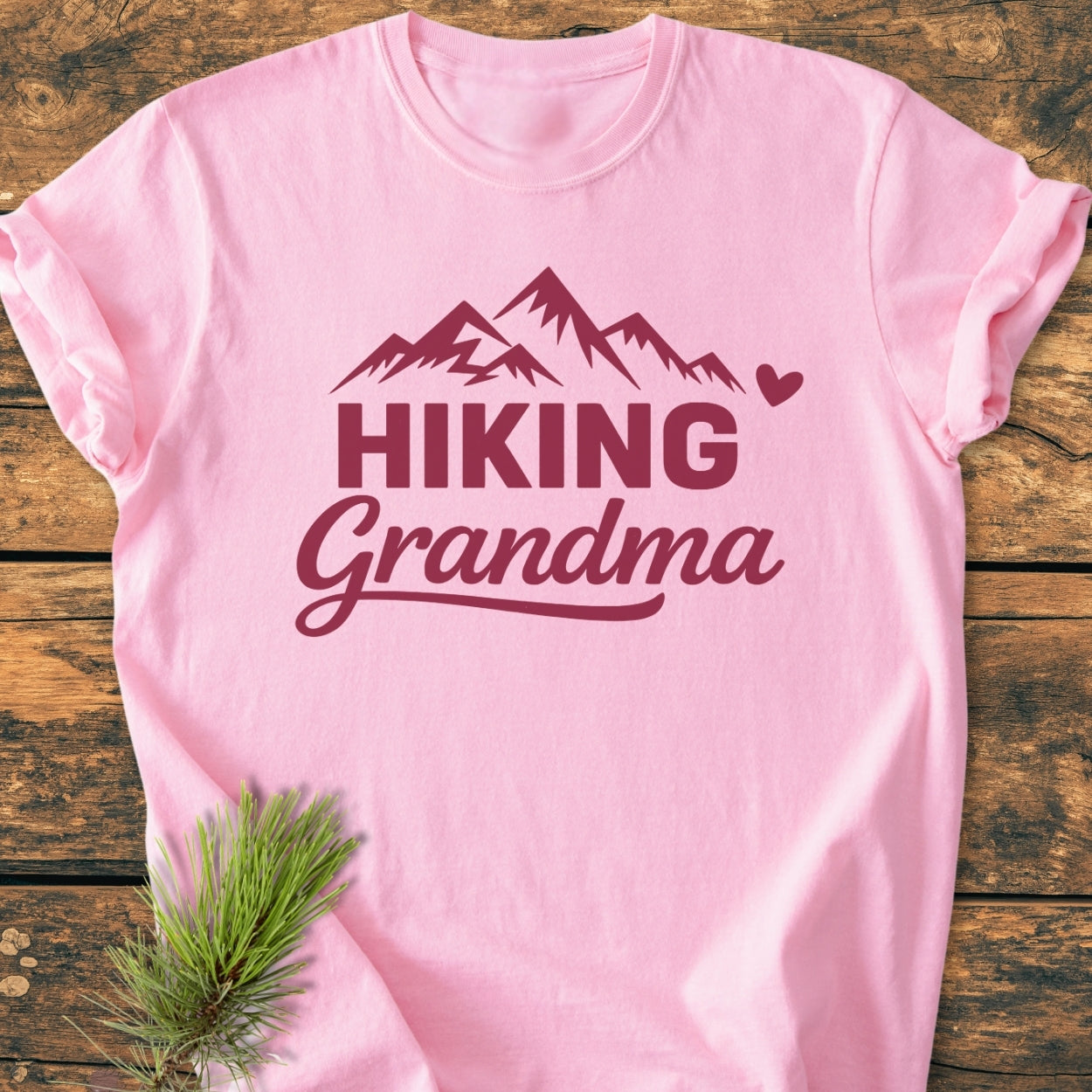 Hiking Grandma - Vintage Wash Tee: Light-pink T-shirt with maroon mountain graphic and HIKING Grandma text, laid flat on wood. Features soft-washed, garment-dyed fabric for comfort, with a pine branch accent nearby.