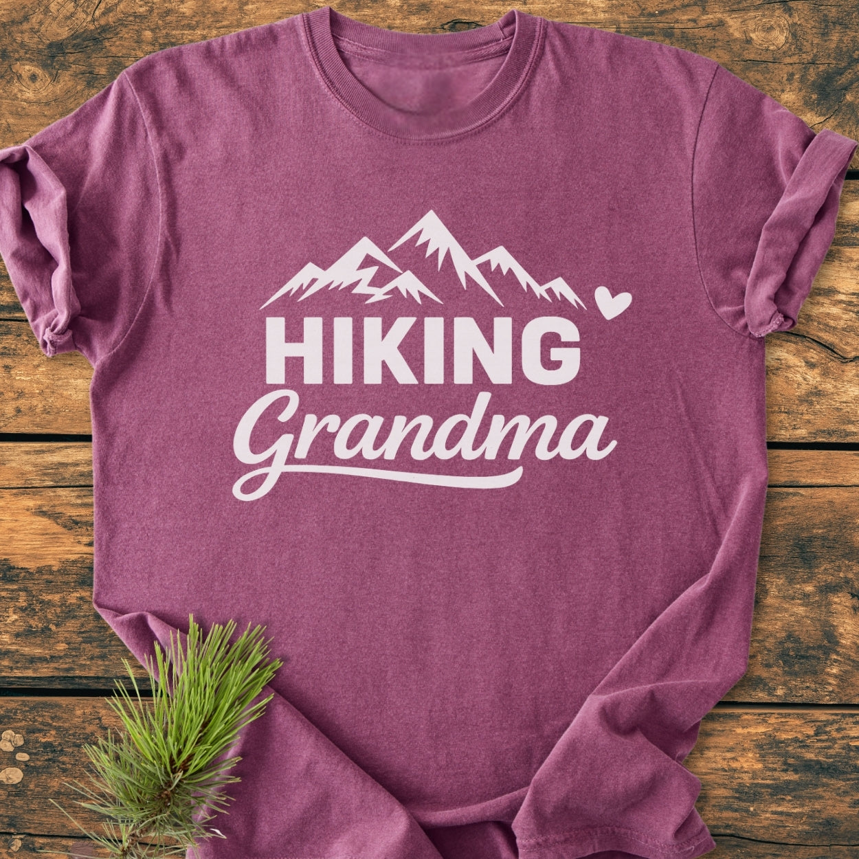 Hiking Grandma - Vintage Wash Tee displayed on wood, featuring a white mountain graphic and text. Includes a heart detail. Pine branch accents the bottom, highlighting the garment's relaxed, soft-washed texture.