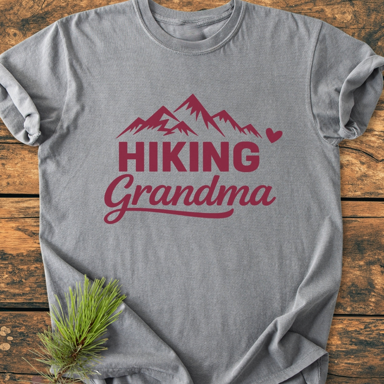 Heather-gray Hiking Grandma - Vintage Wash Tee with deep red mountain print, laid flat on wood. Displayed with a pine sprig, showcasing relaxed fit and durable double-needle stitching.