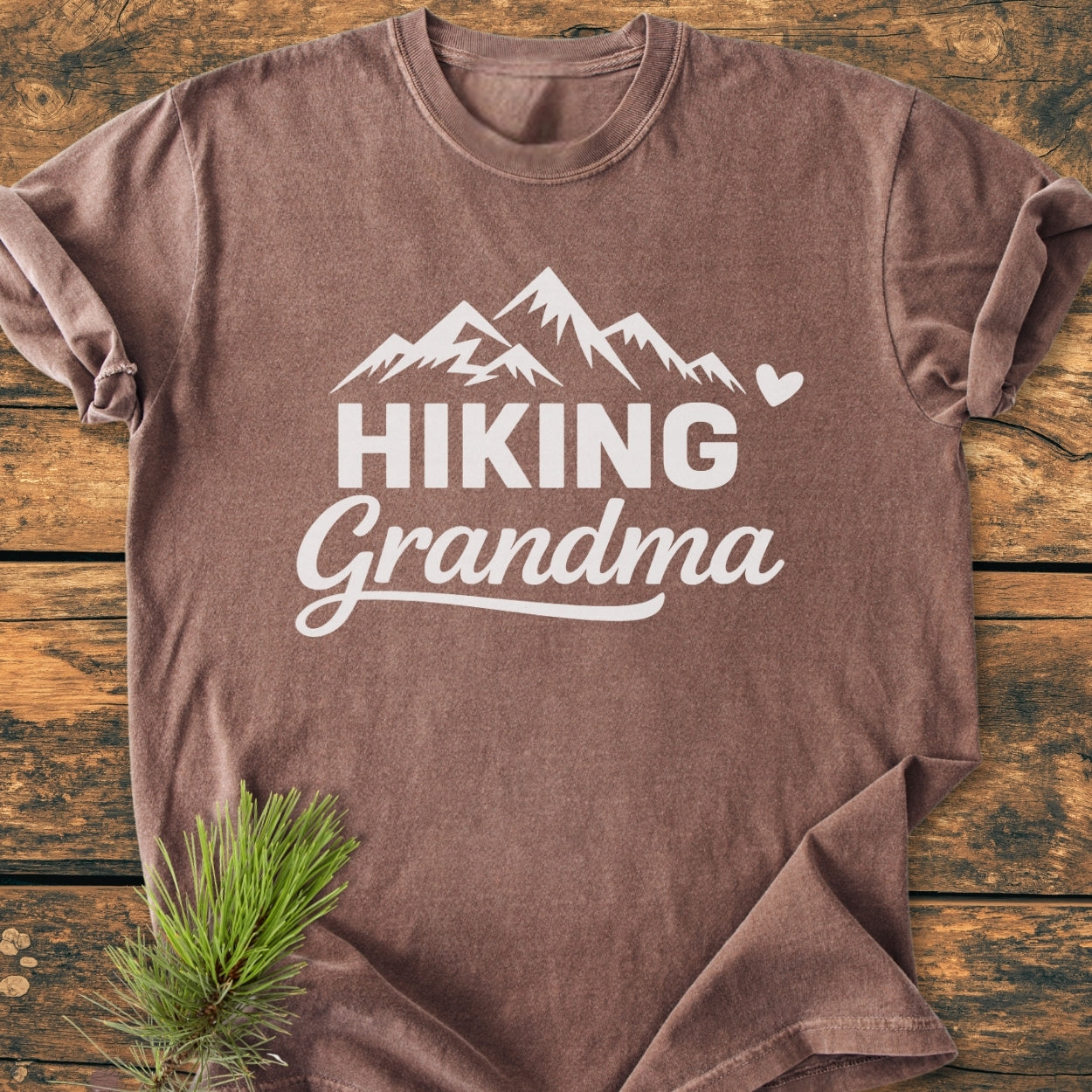 Hiking Grandma - Vintage Wash Tee displayed on a wooden surface, featuring a white mountain graphic and text. The soft-washed, relaxed fit shirt is folded with visible double-needle stitching for durability.