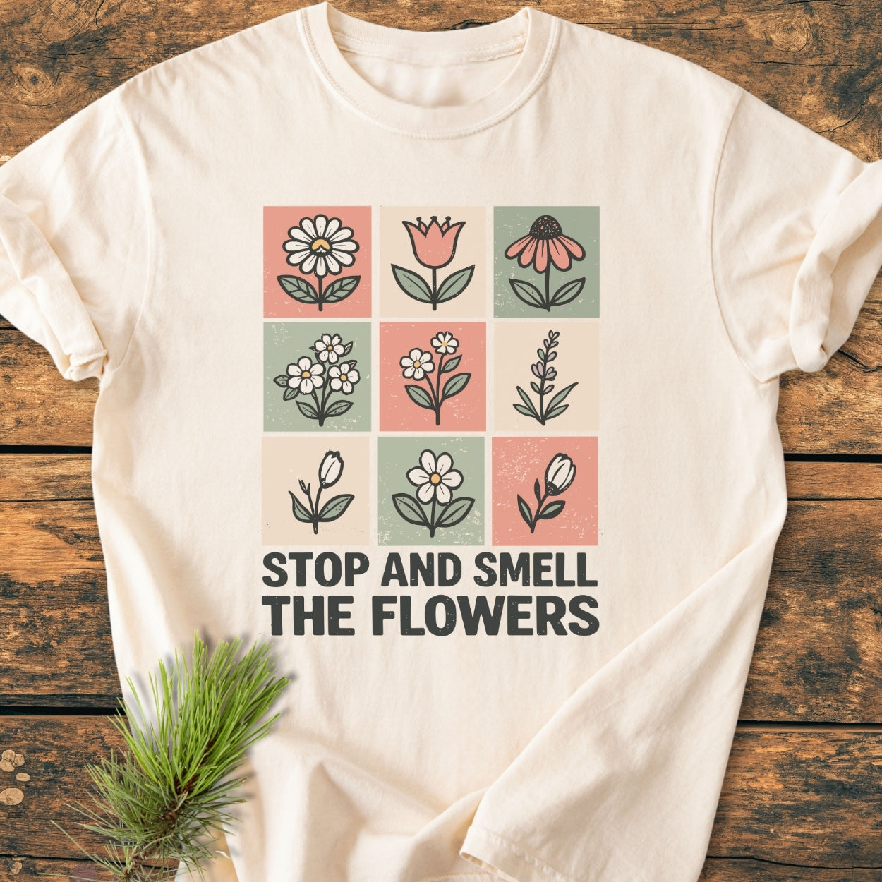 Stop And Smell The Flowers - Vintage Wash Tee: Cream-colored T-shirt featuring a 3x3 grid of hand-drawn flowers in pastel blocks with the slogan, “STOP AND SMELL THE FLOWERS,” laid on wood.