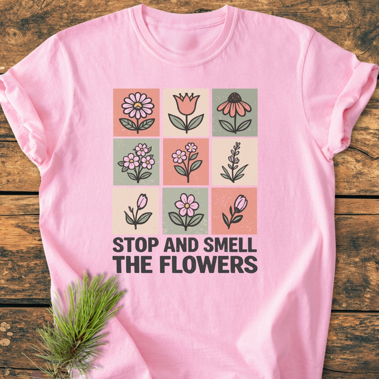 Stop And Smell The Flowers - Vintage Wash Tee featuring a 3×3 grid of hand-drawn flowers and bold slogan on a light pink background, styled with a pine branch on a wooden surface.