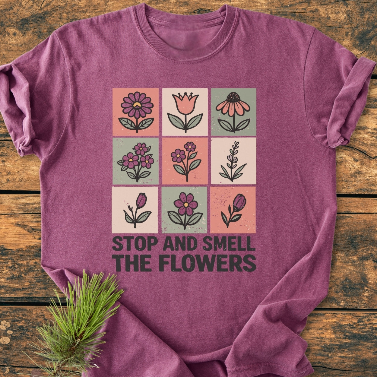Stop And Smell The Flowers - Vintage Wash Tee features a 3x3 grid of hand-drawn flowers, bold text underneath on heather-purple fabric, styled with a pine sprig on a wooden surface.