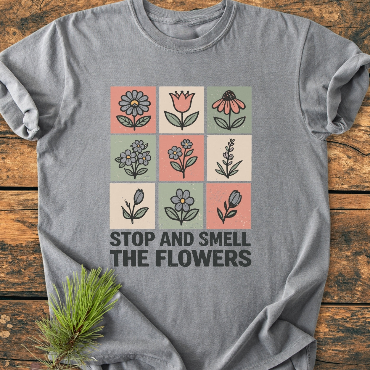 Stop And Smell The Flowers - Vintage Wash Tee: Heather-gray T-shirt featuring a 3x3 grid of pastel flower icons with a bold slogan. Laid on wood with a pine sprig accent.