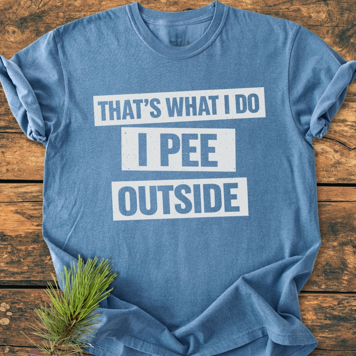 Heather-blue crew-neck T-shirt, That's What I Do. I Pee Outside - Vintage Wash Tee, in white block letters, laid flat on wood. Features a small pine sprig accent.