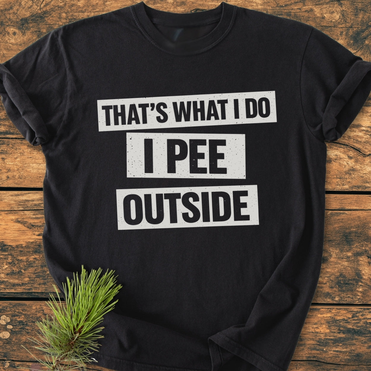 Black T-shirt with “THAT’S WHAT I DO I PEE OUTSIDE” in white letters, laid flat on wood. Part of the Vintage Wash Tee collection, showcasing a relaxed fit and cozy, durable design.