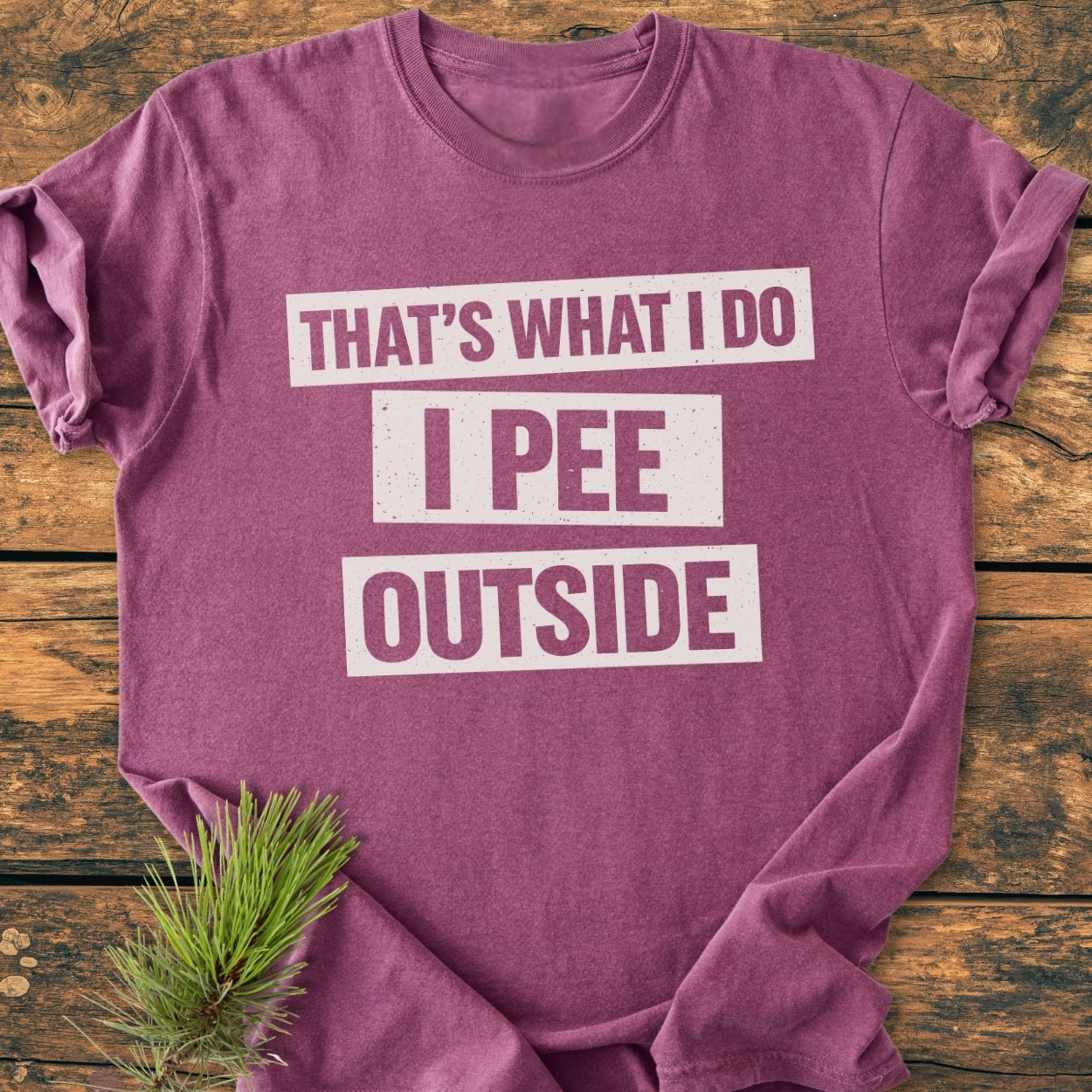 That's What I Do. I Pee Outside - Vintage Wash Tee, displayed folded on wood, features bold white text on plum cotton, styled with pine needles; soft, durable, relaxed fit for daily wear.