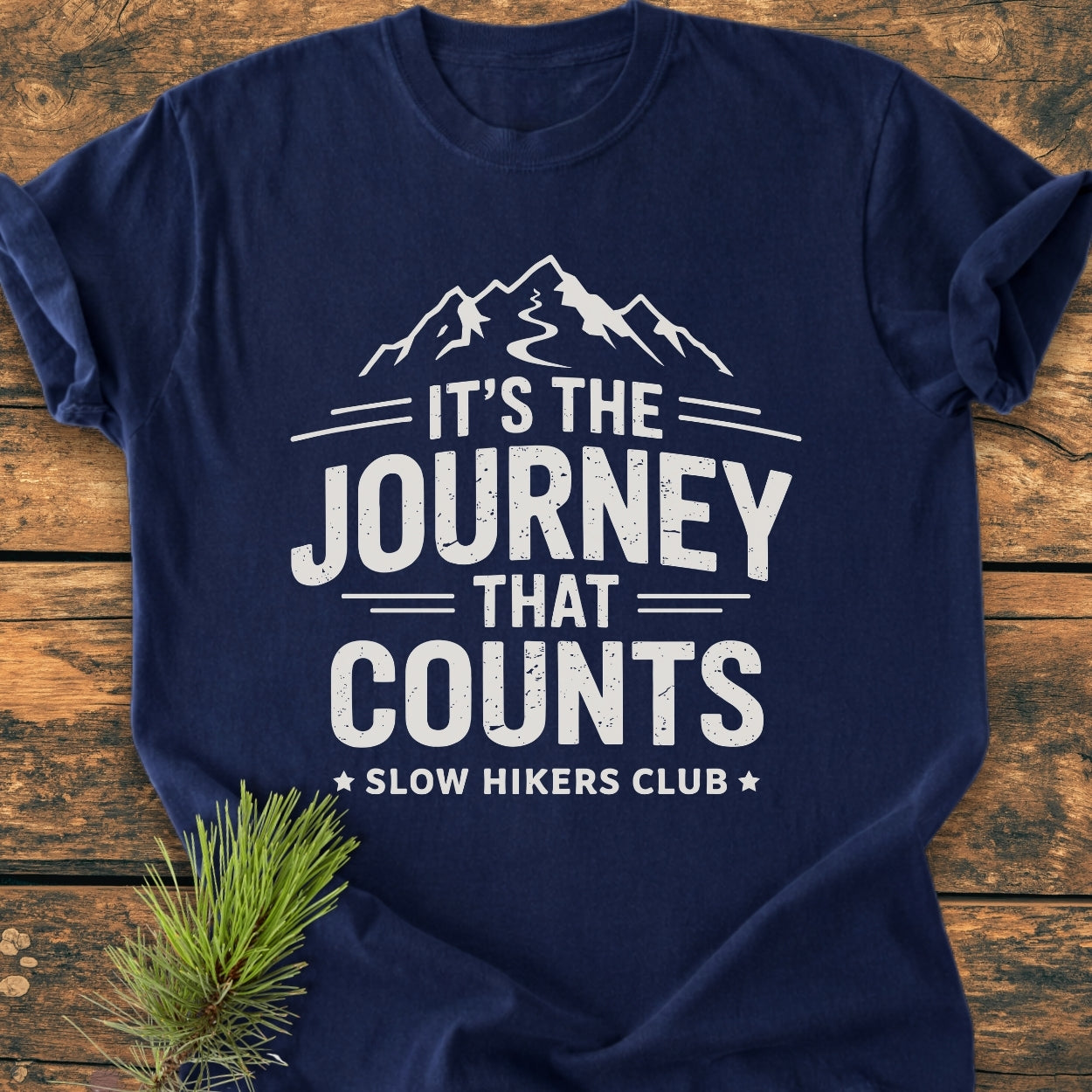 It's The Journey That Counts - Vintage Wash Tee, featuring stylized mountain peaks and slogans, laid on a wooden surface with a pine sprig; crafted from soft, durable, garment-dyed cotton for everyday comfort.