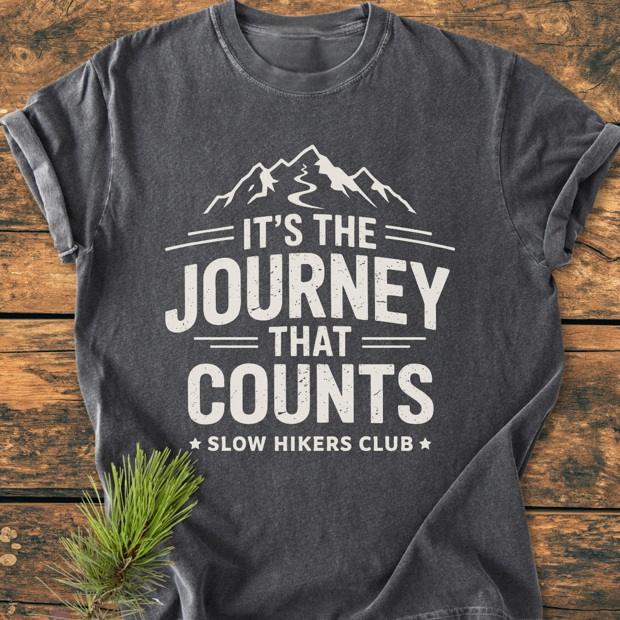 Dark-gray It's The Journey That Counts - Vintage Wash Tee featuring a mountain graphic and Slow Hikers Club text, laid flat on a rustic wooden surface with a pine sprig for styling.