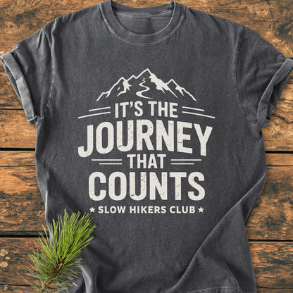Dark-gray It's The Journey That Counts - Vintage Wash Tee featuring a mountain graphic and Slow Hikers Club text, laid flat on a rustic wooden surface with a pine sprig for styling.