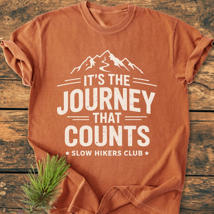 It's The Journey That Counts - Vintage Wash Tee features a white mountain graphic and slogan on a rust-orange shirt, symbolizing outdoor adventures. Laid flat with pine needles, exudes a hiking theme.