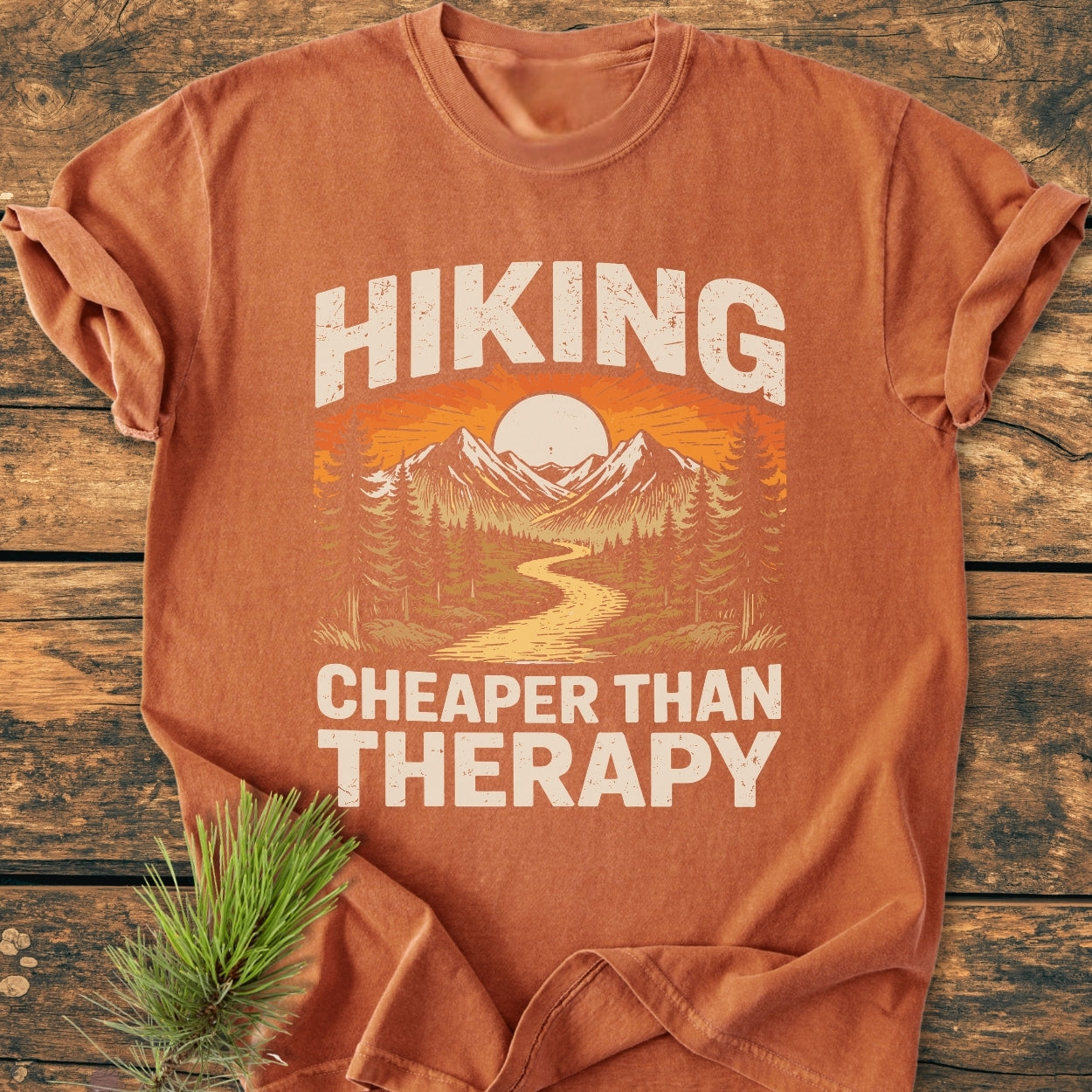 Hiking Cheaper Than Therapy - Vintage Wash Tee, featuring a stylized outdoor scene with mountains and sun, laid on wood. The fabric is soft-washed, 100% ring-spun cotton for comfort and durability.