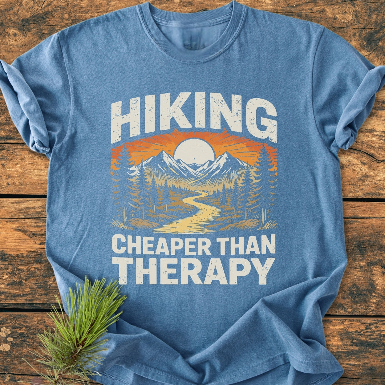 Heather-blue crew-neck T-shirt with a stylized outdoor scene and the words HIKING and CHEAPER THAN THERAPY, placed on a wooden surface, embodying the Hiking Cheaper Than Therapy - Vintage Wash Tee.