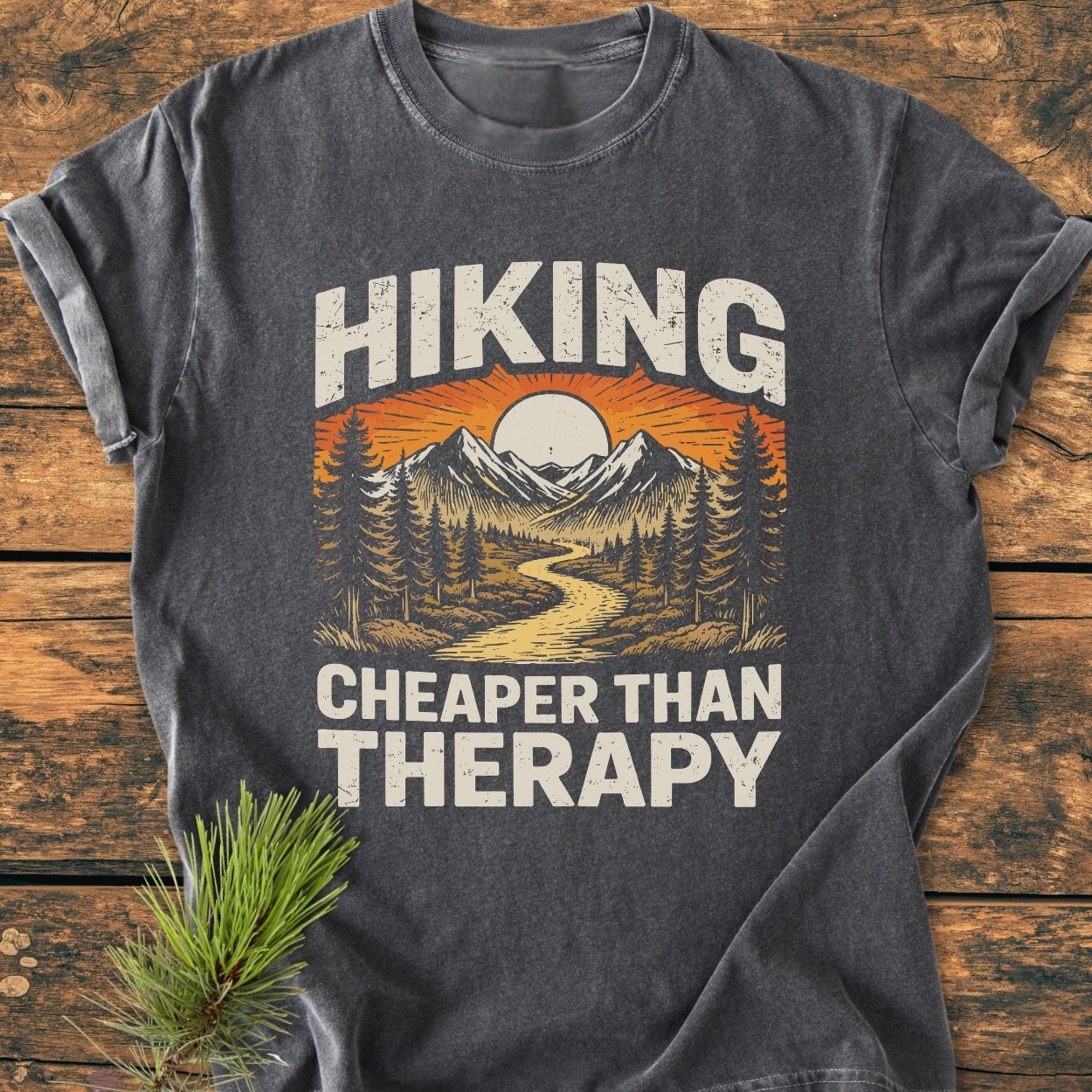 Hiking Cheaper Than Therapy - Vintage Wash Tee features a stylized outdoor scene with mountains and a sun, emphasizing adventure. Laid on wood, it highlights its relaxed fit and durable design.