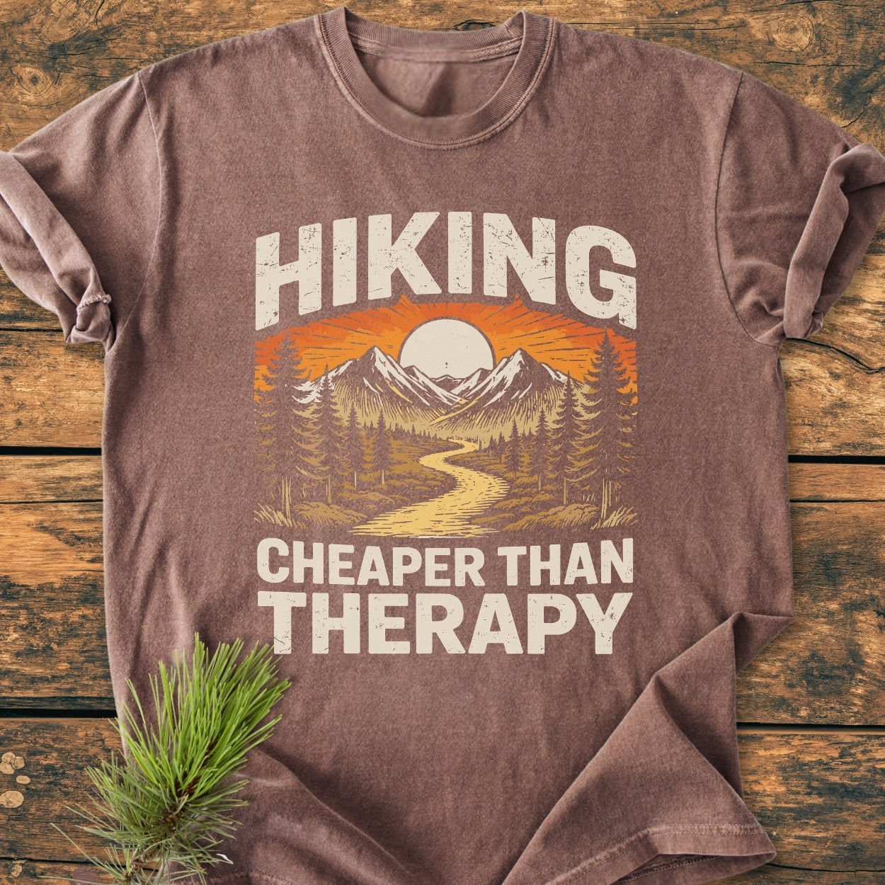 Hiking Cheaper Than Therapy - Vintage Wash Tee features a stylized outdoor scene with mountains and sun, laid flat on wood. A pine sprig accents this brown, soft-washed, relaxed-fit cotton T-shirt.