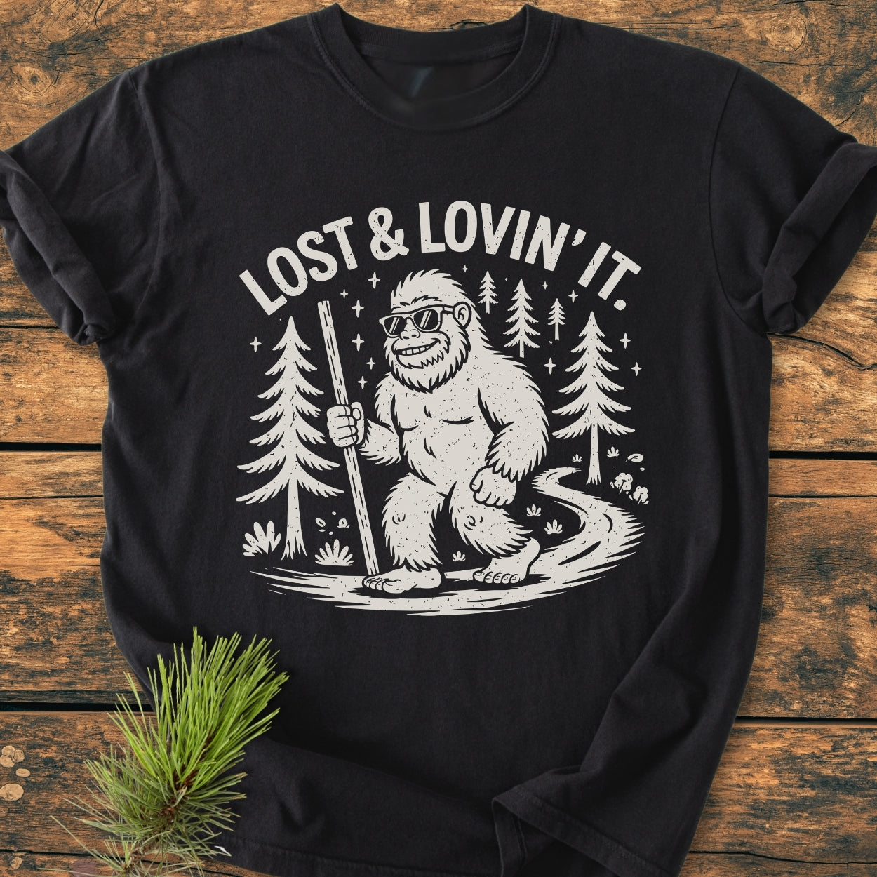Lost And Lovin' It - Vintage Wash Tee features a cartoon Sasquatch with sunglasses and hiking stick, surrounded by forest elements, slogan above; displayed on rustic wood, highlighting its cozy, relaxed fit.