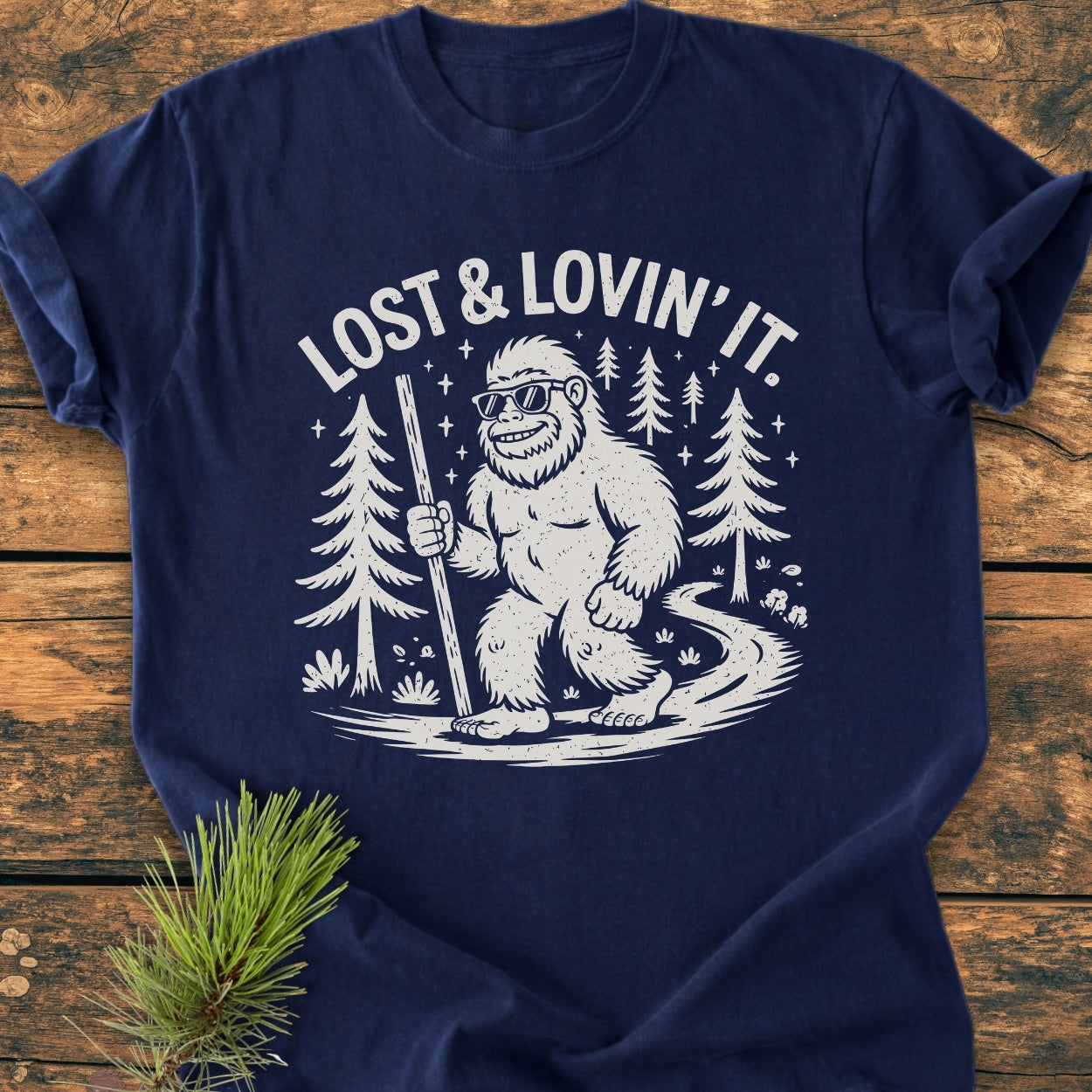Lost And Lovin' It - Vintage Wash Tee features a cartoon Sasquatch with sunglasses on a forest path, slogan above. Made of soft, durable 100% ring-spun cotton for daily comfort.