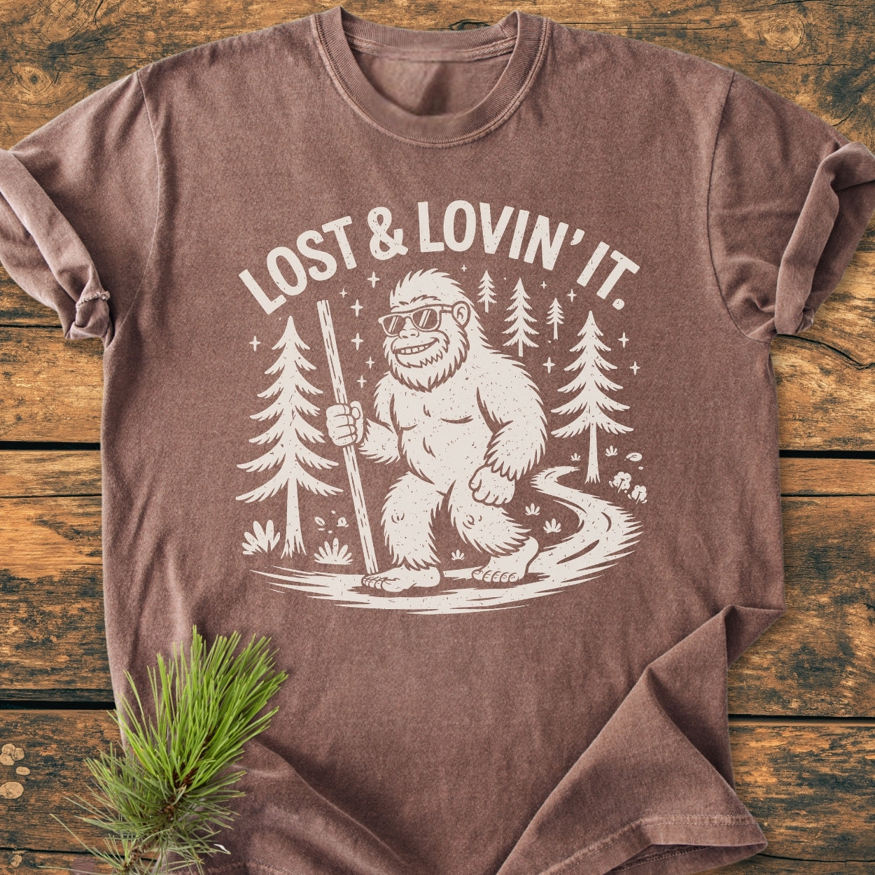 Lost And Lovin' It - Vintage Wash Tee featuring a cartoon Sasquatch with sunglasses, hiking on a forest path, printed on a soft brown Comfort Colors garment-dyed T-shirt with a pine bough accent.