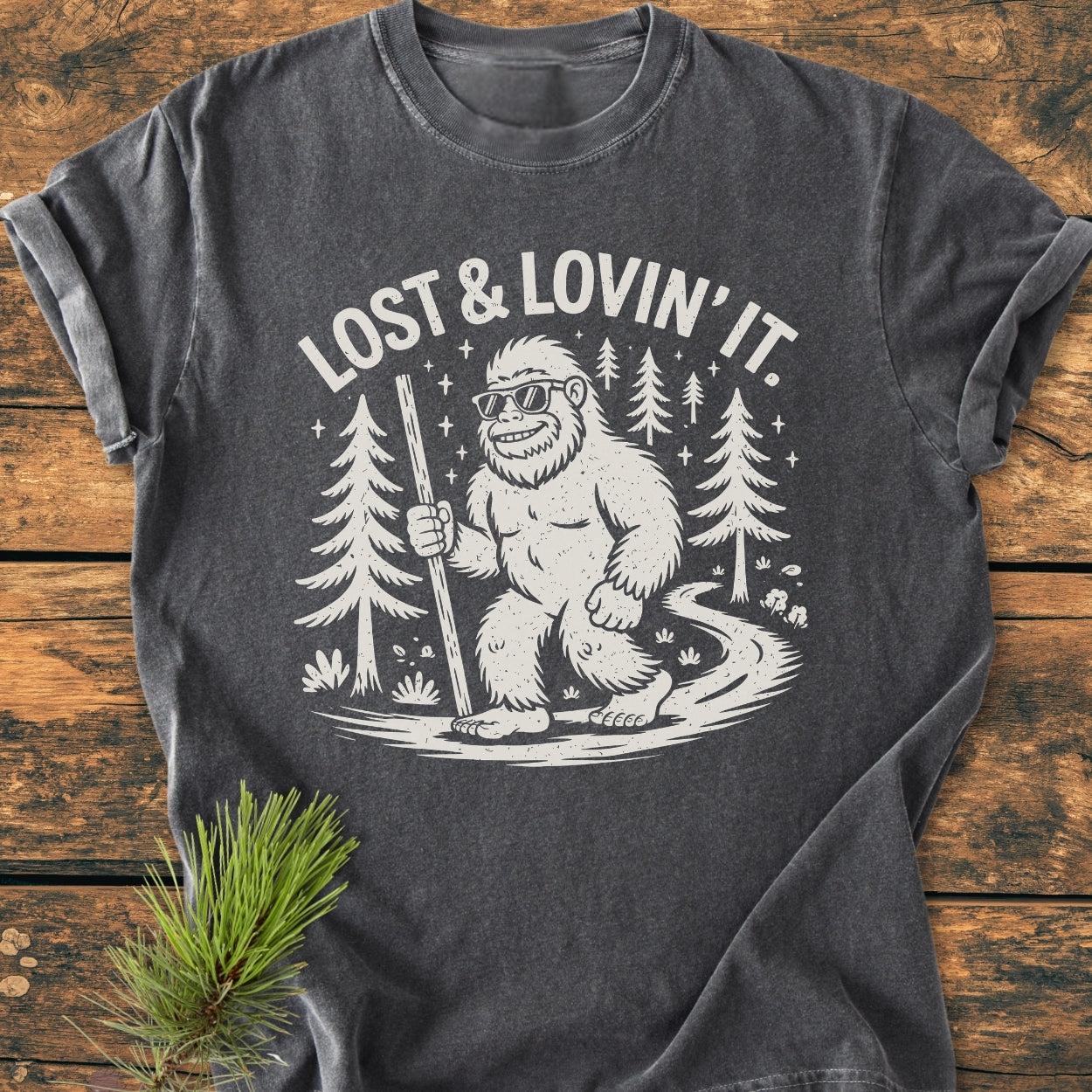 Lost And Lovin' It - Vintage Wash Tee featuring a cartoon Sasquatch in sunglasses with a hiking stick on a forest path, printed on a dark-grey T-shirt styled with a pine sprig.