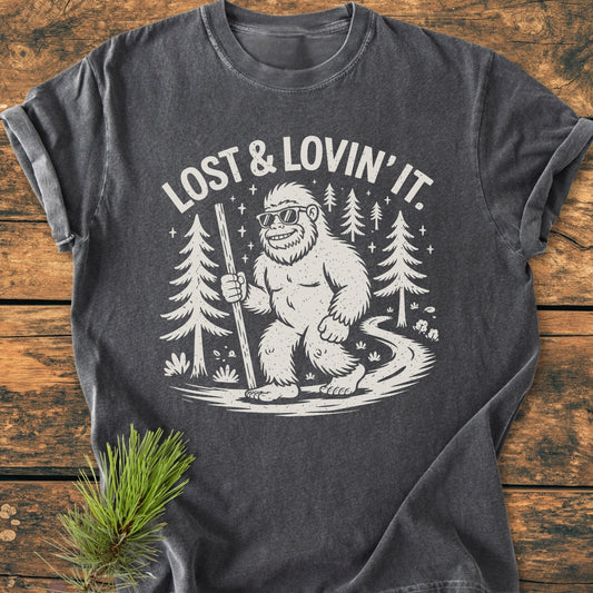 Lost And Lovin' It - Vintage Wash Tee featuring a cartoon Sasquatch in sunglasses with a hiking stick on a forest path, printed on a dark-grey T-shirt styled with a pine sprig.