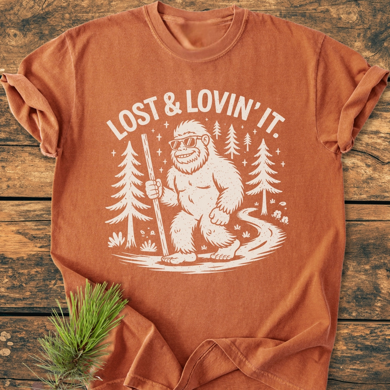 Lost And Lovin' It - Vintage Wash Tee featuring a cartoon Bigfoot with sunglasses, walking stick, and forest path design. Laid on wood surface, with pine branch accentuating its rustic charm.