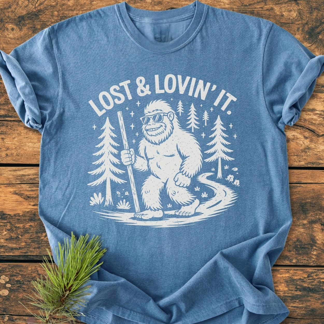 Lost And Lovin' It - Vintage Wash Tee featuring a cartoon Bigfoot with sunglasses on a forest trail, slogan above. Heather-blue, soft-washed, 100% ring-spun cotton, relaxed fit, durable stitching.