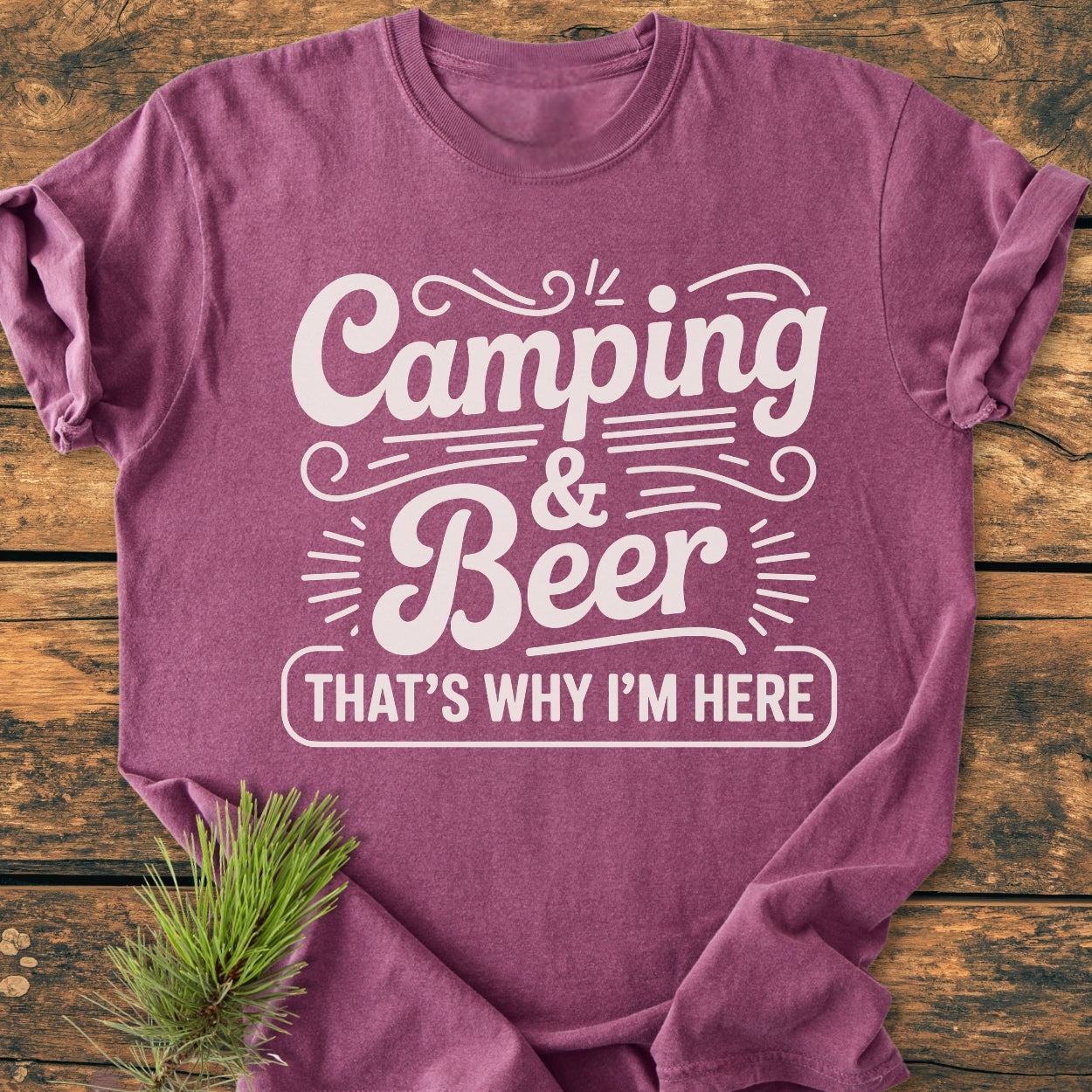 Camping And Beer - Vintage Wash Tee folded on a wooden surface, featuring white stylized lettering. A small pine branch accentuates the shirt's heathered appearance, emphasizing its cozy, relaxed fit.