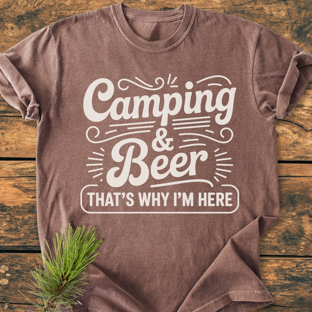 The Camping And Beer - Vintage Wash Tee, a brown short-sleeve T-shirt, features a white slogan, Camping & Beer That's Why I'm Here, laid on rustic wood with a pine sprig accent.