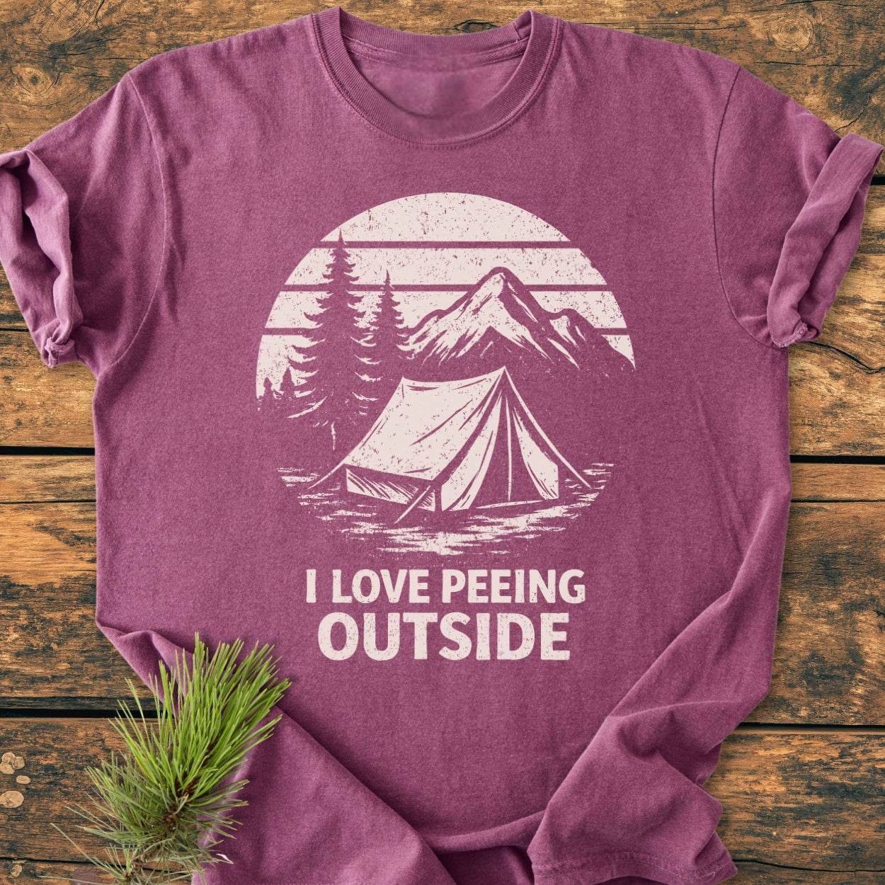 I LOVE Peeing Outside - Vintage Wash Tee features a maroon-purple crewneck with a tent and pine trees print, laid on wood with a pine branch accent, offering comfort and durability.