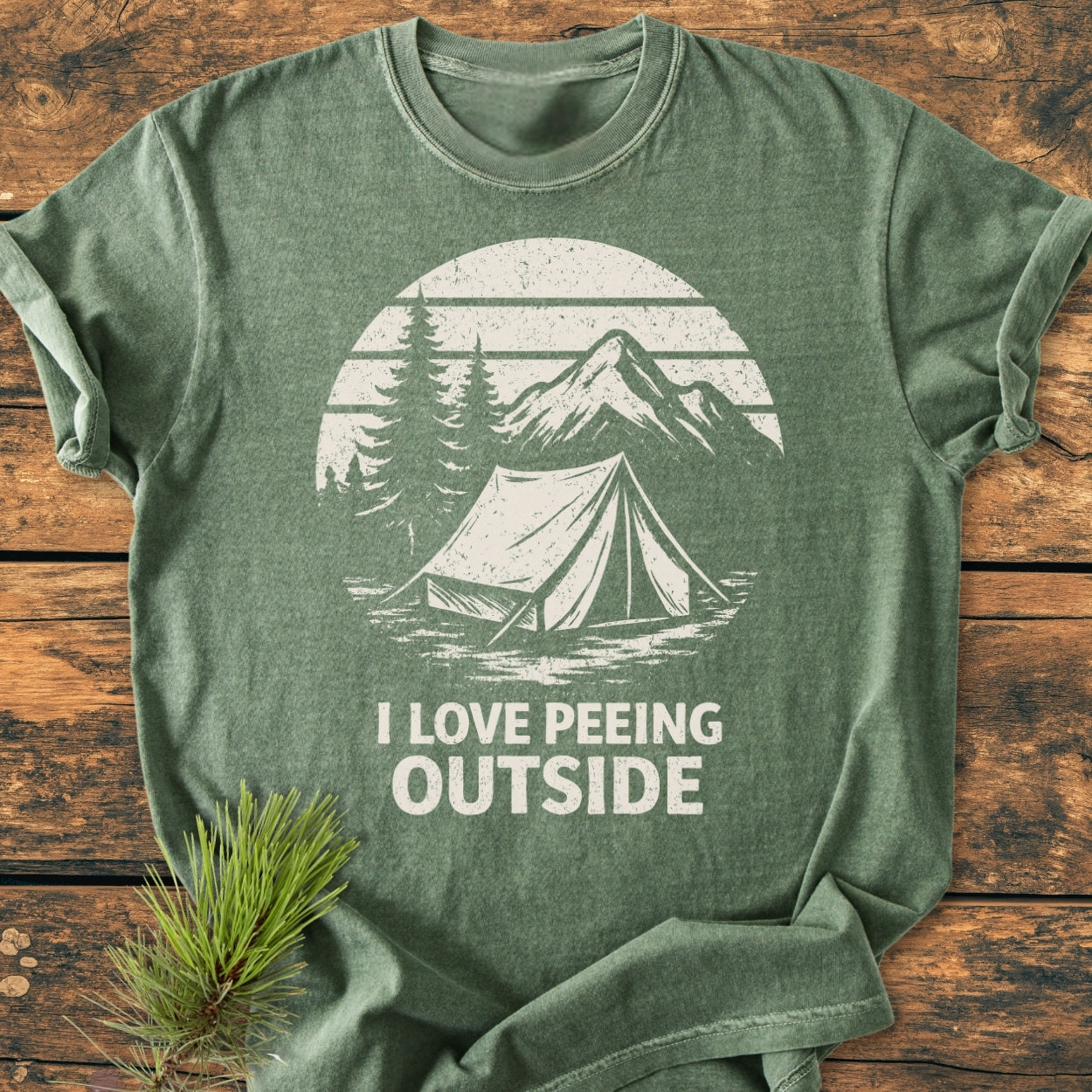Heather-green I LOVE Peeing Outside - Vintage Wash Tee features a distressed camping scene graphic with a tent, pine trees, and mountain, evoking outdoor humor. Displayed on a wooden surface with pine sprig.