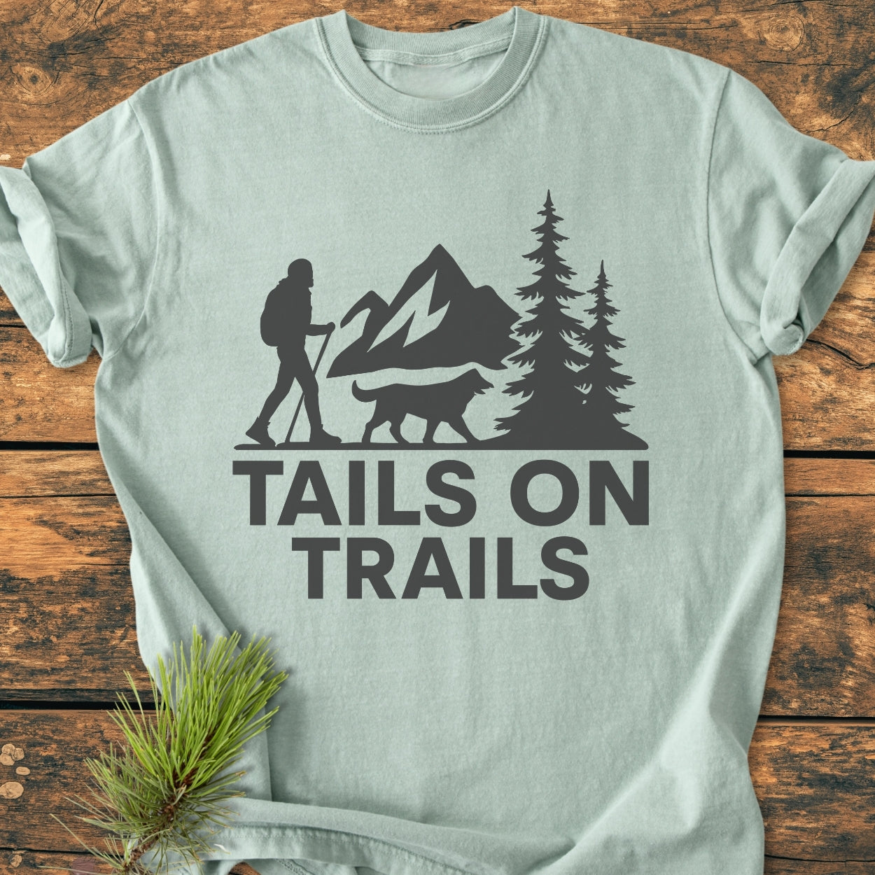 Tails On Trails - Vintage Wash Tee featuring a nature-themed logo of a hiker and dog with mountains and trees, displayed on a wooden surface with a pine sprig for styling.