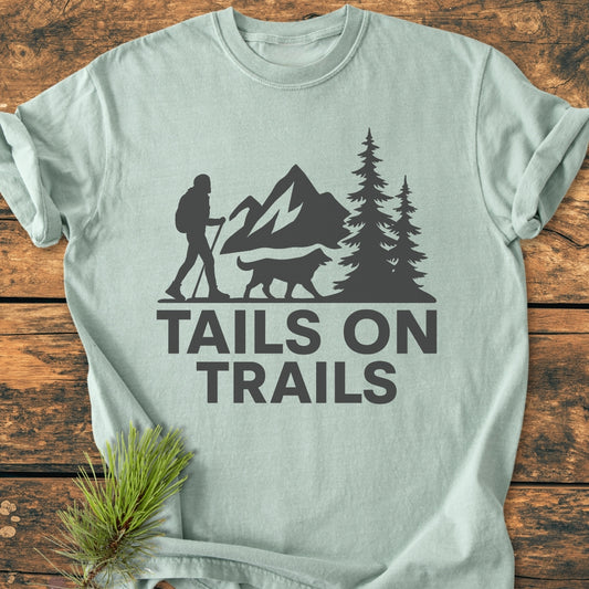 Tails On Trails - Vintage Wash Tee featuring a nature-themed logo of a hiker and dog with mountains and trees, displayed on a wooden surface with a pine sprig for styling.