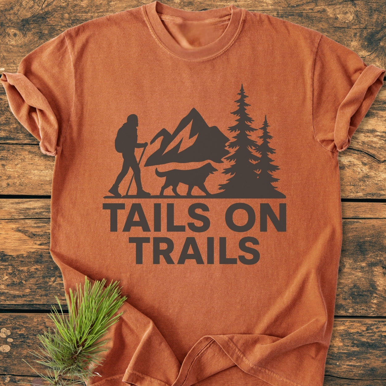 Tails On Trails - Vintage Wash Tee features a hiker and dog silhouette against mountains, printed on rust-orange garment-dyed fabric, laid flat with pine styling, highlighting its outdoor adventure theme.
