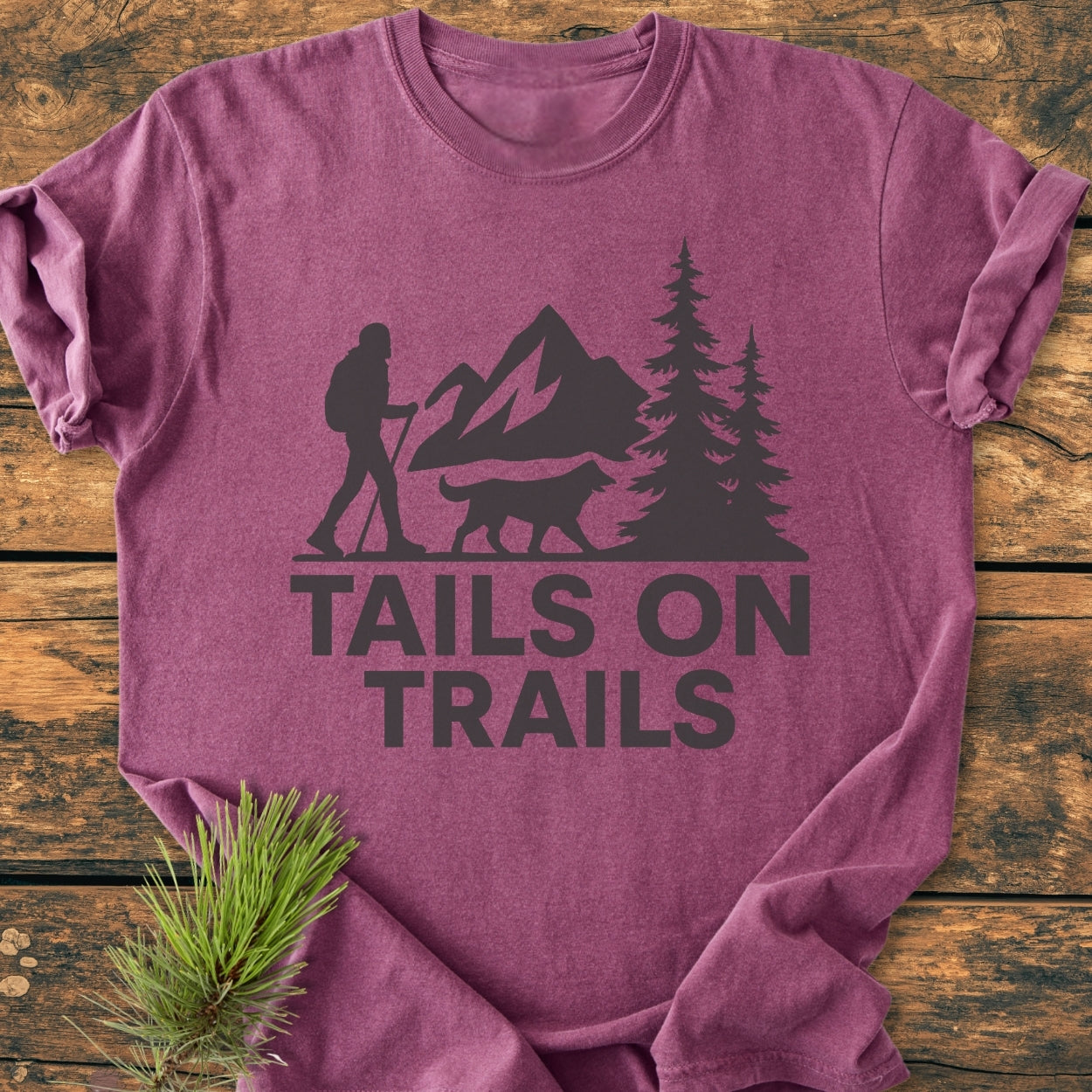 Tails On Trails - Vintage Wash Tee features a hiker and dog silhouette with mountain peaks and pine trees, printed on a heathered-purple t-shirt, laid on wood, accompanied by a pine branch.