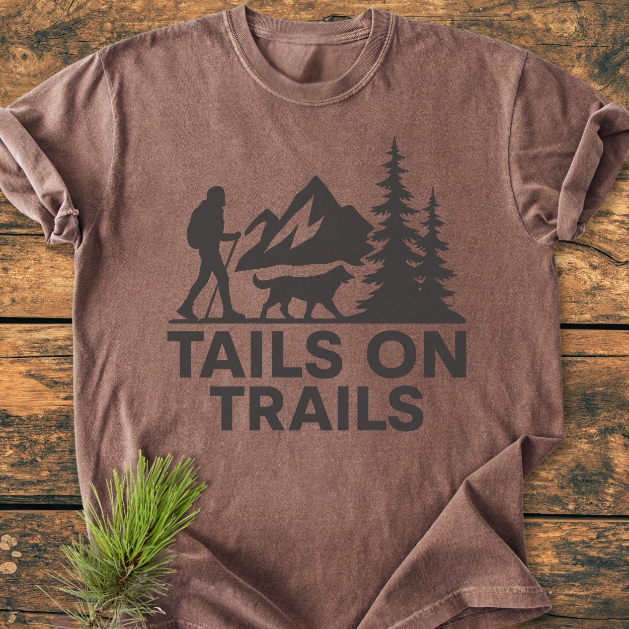 Tails On Trails - Vintage Wash Tee featuring a hiker and dog silhouette with mountain scene, printed in black on a soft, folded brown-heather T-shirt, laid on a rustic wooden backdrop.