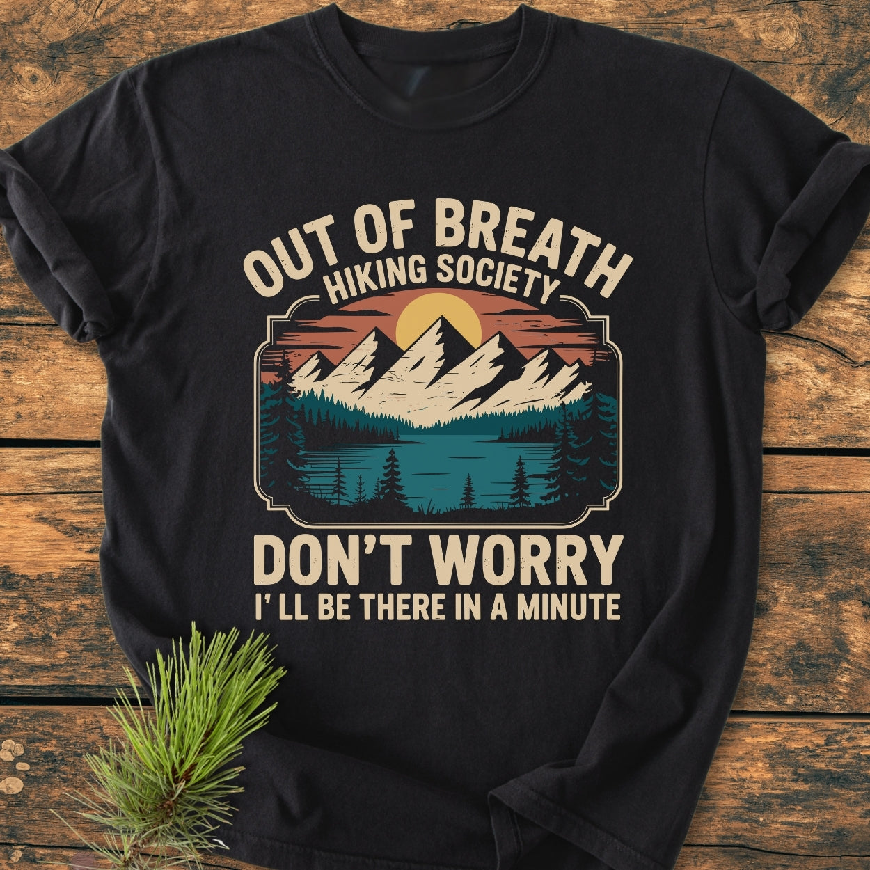 Out Of Breath Hiking Society - Vintage Wash Tee, featuring a retro sun-mountain emblem and humorous text, laid on wood. Made of soft, durable 100% ring-spun cotton, offering a relaxed fit.