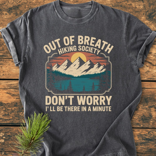 Out Of Breath Hiking Society - Vintage Wash Tee featuring a retro nature graphic with mountains, trees, and a lake, humorously captioned, laid flat on a rustic wood background.