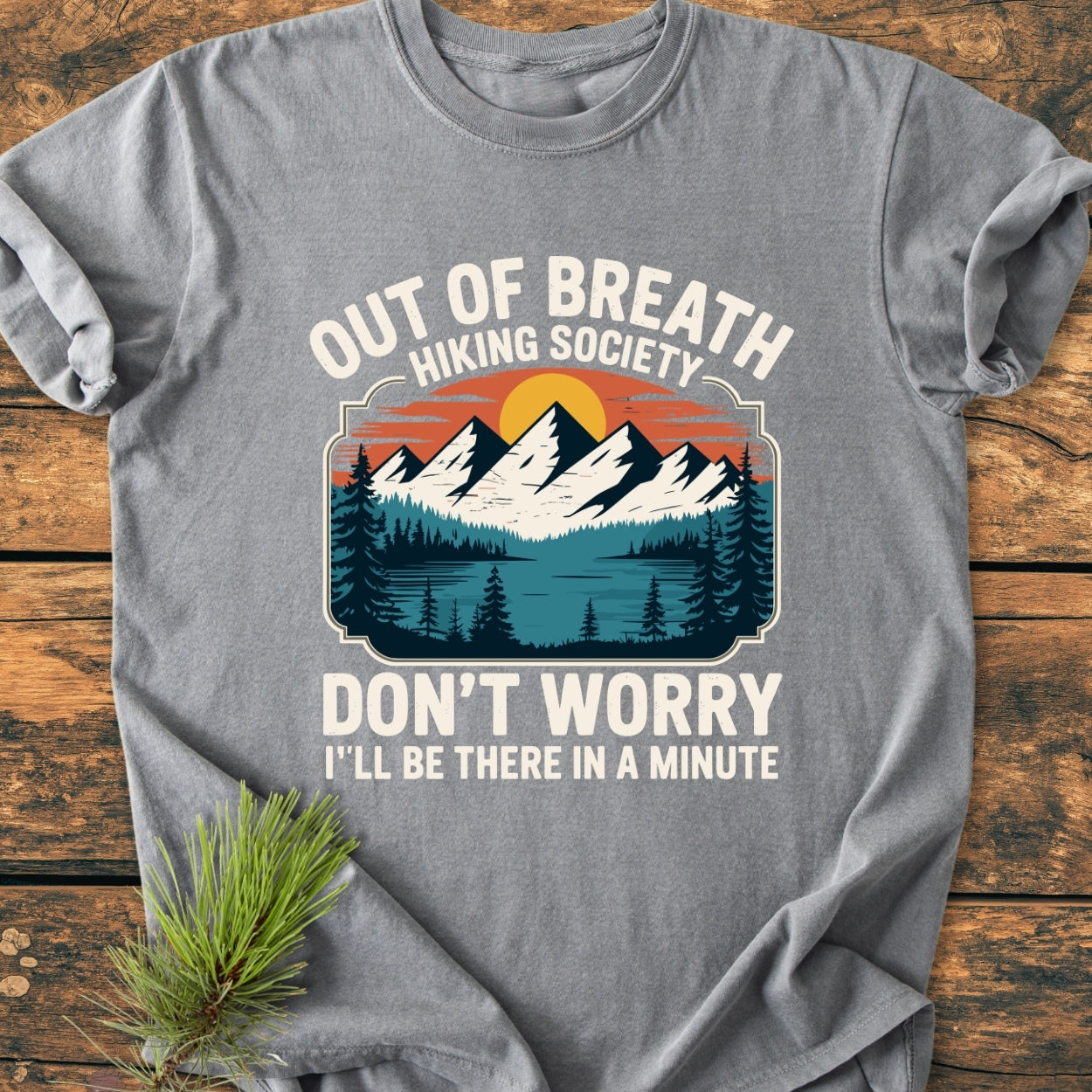 Out Of Breath Hiking Society - Vintage Wash Tee on rustic wood, features a stylized outdoor scene with mountains, trees, and sun. Includes humorous text above and below the graphic.