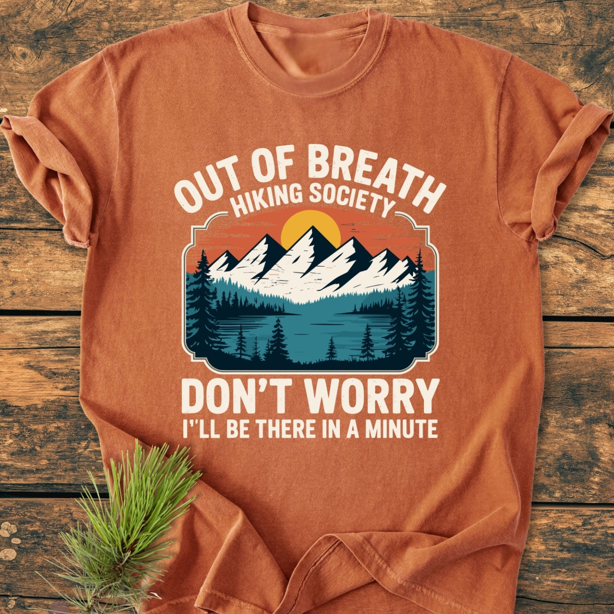 Out Of Breath Hiking Society - Vintage Wash Tee: Orange-rust t-shirt features a retro camp graphic with mountains and a lake, surrounded by pine trees, perfect for hiking enthusiasts. Laid on a wooden surface.