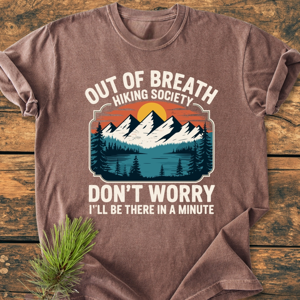 Out Of Breath Hiking Society - Vintage Wash Tee featuring a retro mountain scene and humorous text, displayed on a rustic surface with a pine sprig, showcasing its relaxed, garment-dyed cotton fabric.