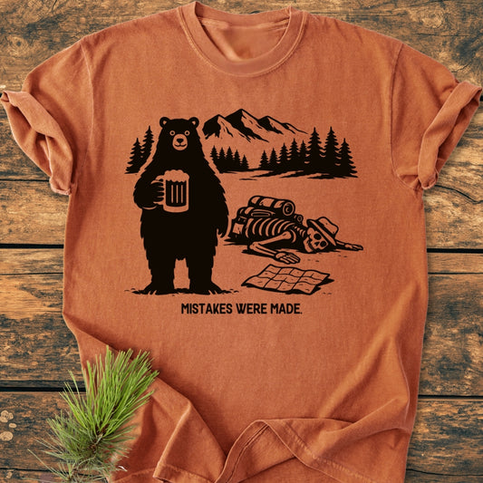Mistakes Were Made - Vintage Wash Tee features a comic-style bear holding a beer and a skeleton with a map, set against a mountain landscape. The tee is laid on a wooden surface.