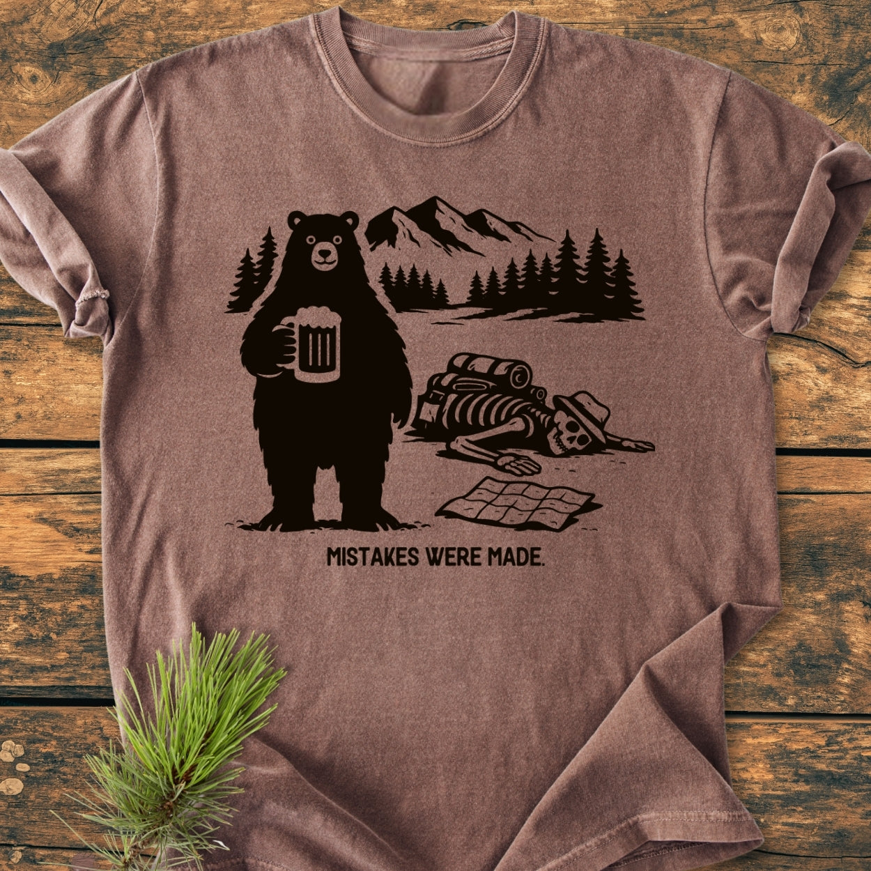 Mistakes Were Made - Vintage Wash Tee features a bear with beer and a hiker’s skeleton beside a caption. Pine trees and mountains form the backdrop, enhancing the shirt's quirky design.