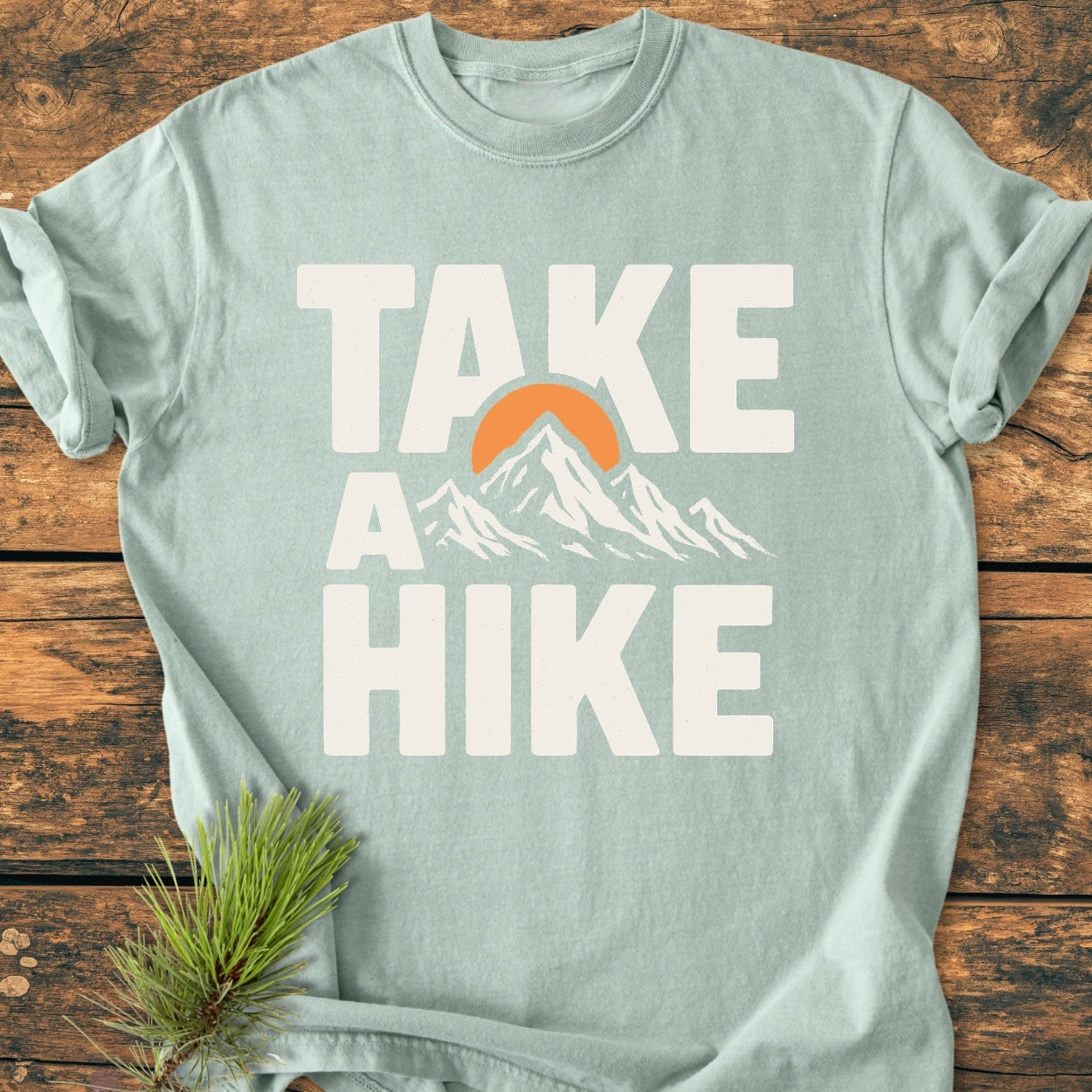 Take A Hike - Vintage Wash Tee in seafoam, featuring a bold graphic with “TAKE A HIKE” text, mountain silhouette, and orange sun. Laid on a wooden surface with a pine sprig decoration.
