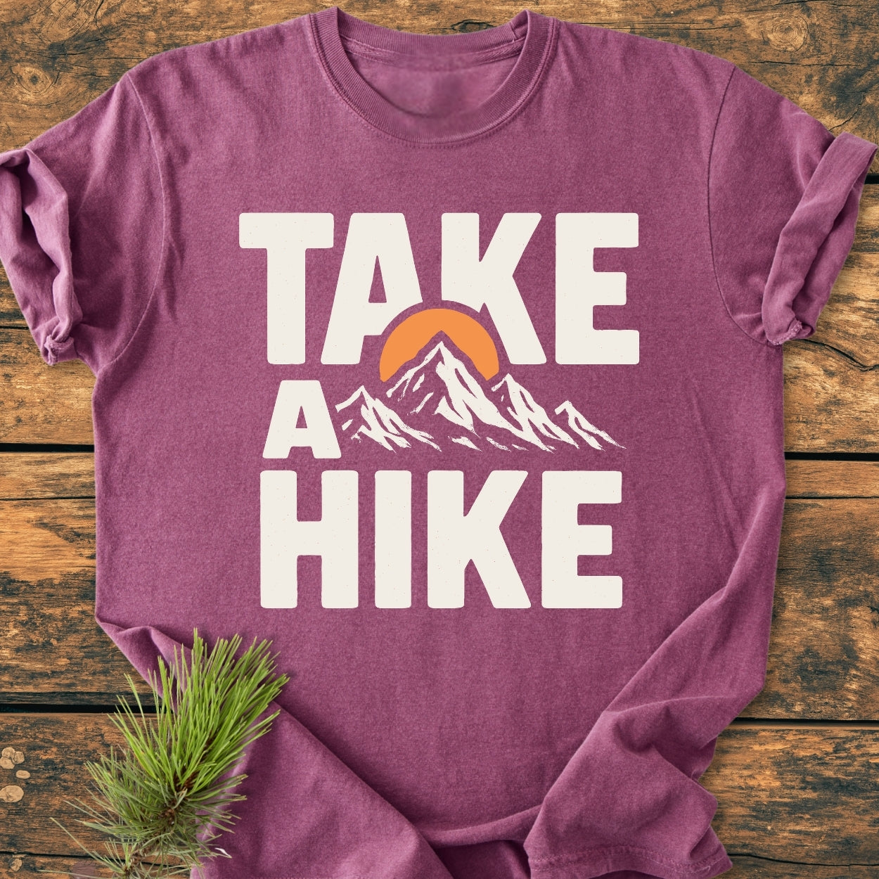 Take A Hike - Vintage Wash Tee features a heathered purple shirt with a bold white slogan and mountain design, styled with a pine sprig on rustic wood, embodying an outdoor spirit.
